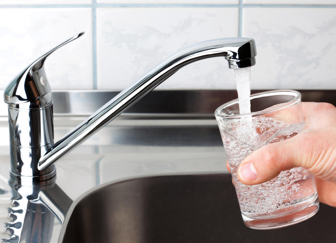 Eine Hand mit einem Wasserglas rechts im Bild, links ein geöffneter Wasserhahn, aus dem Leitungswasser ins Glas fließt.